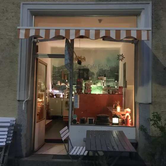 Das Café von außen in der Dämmerung, die Fenster beleuchtet, davor Tisch und Bank