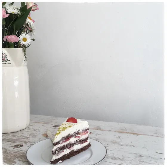 Ein Stück Sahnetorte mit Schokoraspeln und einer Kirsche, dahinter eine Vase mit Blumen